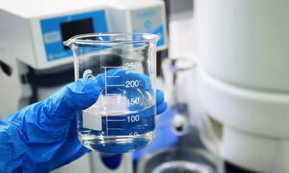 Blue-gloved hand holding a glass beaker of clear liquid in a laboratory