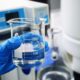 Blue-gloved hand holding a glass beaker of clear liquid in a laboratory