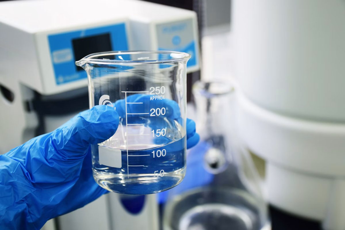 Blue-gloved hand holding a glass beaker of clear liquid in a laboratory